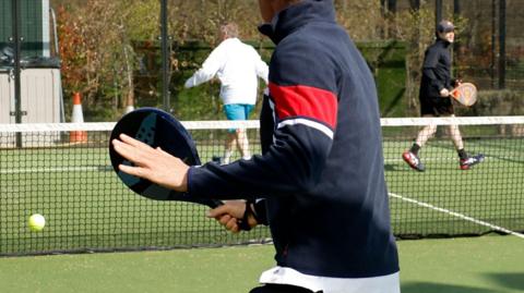 A man wearing a hoodie prepares to play a shot with a padel racquet as the ball comes towards him. In the background, two figures, one of whom is also holding a racquet, walk on the other side of the net.