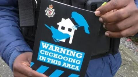 A close up of a police officer wearing a protection vest and long sleeved blue raincoat as he holds a poster saying "Warning cuckooing in this area". The poster has the Avon and Somerset Police branding on it, and a house with two blue cuckoo outlines on it.