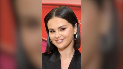 Selena Gomez has dark hair in a bob. She wears a black jacket and hoop earrings.