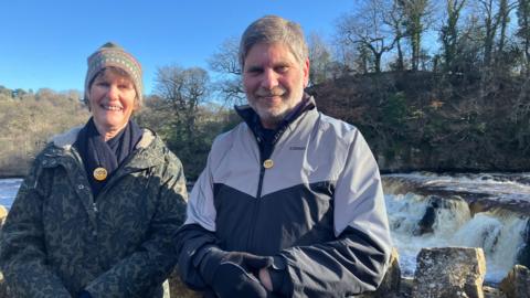 A woman in a green jacket and a scarf and hat stood next to a man in a grey and navy jacket and gloves. Both are wearing yellow badges with SOS written on them. There is a river behind them and water can be seen flowing down a small waterfall. Trees in the background.