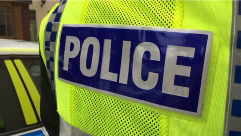 The back of a British police vest. It is bright yellow with a plastic blue rectangle logo in the middle with the text POLICE in white lettering