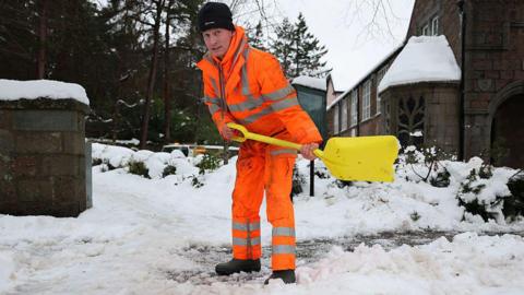A man in a orange high-vis suit shovels snow.