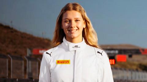 Rachel Robertson wearing a white racing suit with various sponsors logos on it. The metal fencing of a racetrack can be seen in the background.