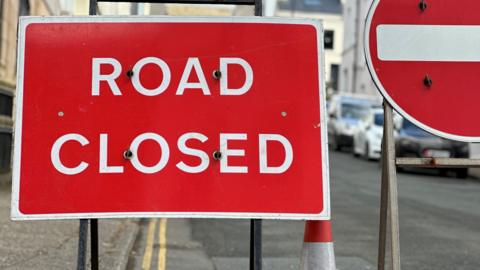 A Road Closed sign, which is red with white writing. A section of a circular red and white No Entry sign is to the right of it.