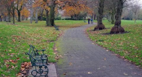 A gravel path winds through a park past a green metal bench. There are fallen leaves on the ground and several trees. In the distance, two men - one of them pushing a bike - are walking along the path.