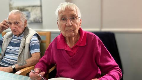 Lilian in a pink sweater eating food and looking at the camera with a serious expression