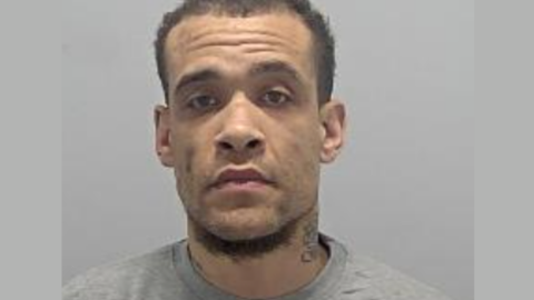 A police mug shot of Callun Bermingham (tbc sp). He has short black hair and a light beard. He is wearing a grey sweatshirt.