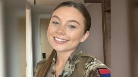 A selfie of Jaysley Beck wearing her military uniform, with her long brown hair slicked back into a low ponytail. She has blue eyes and is smiling at the camera.