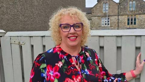 Karen Dickinson with blond hair and purple glasses wearing a black dress with red and violet flowers standing by a grey gate.