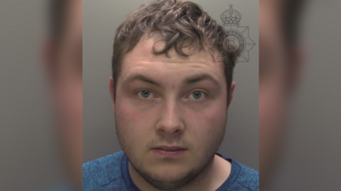 A police custody photo of a man - Dylan Lowther - looking at the camera. He has short brown hair and short brown facial hair. He is wearing a blue top. The wall behind him is grey.