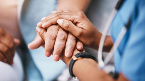 A stock image of a nurse holding the hand of a patient. The nurse is wearing a black watch, and her blue uniform is blurred to the right.
