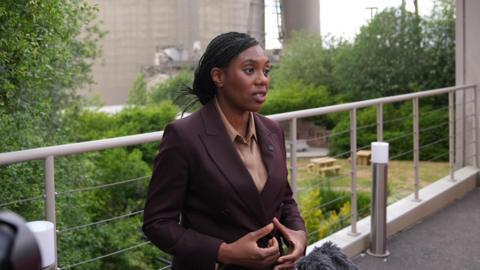 The Conservative leader Kemi Badenoch stands in front of a silo at the Cemex cement plant in Rugby. She is wearing a plum-coloured suit over an light brown blouse.