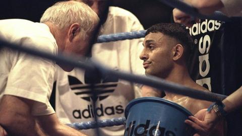 A boxer sits in the corner of a boxing ring, being attended to by his team - one holding an Adidas-branded bucket and others dressed in Adidas gear - offering support and guidance between rounds.