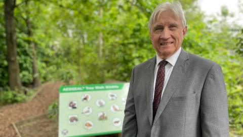 A smartly dressed man in a grey suit and white hair smiles surrounded by woodland. Behind him a green plaque showcases pcitures of woodland animals