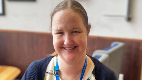 Head-and-shoulders image of a smiling Catherine Makin. She has long brown hair tied back in a ponytail with a centre parting. Smiling for the camera, she is wearing a cream top with a navy cardigan on top.