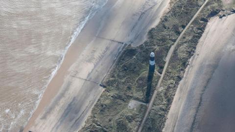 A drone view of a black-and-white lighthouse on a narrow spit of land with green dunes in the centre and sandy beaches and sea water to either side.