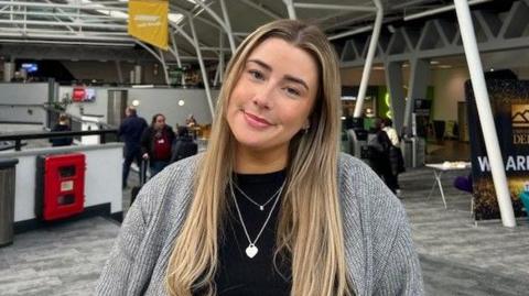 Picture of a white, long haired, blonde, female wearing a grey cardigan with a black top underneath. 