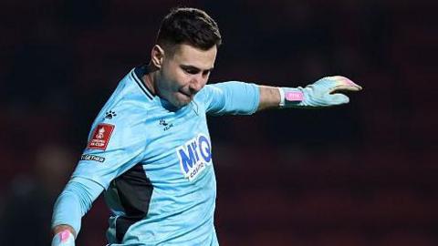 Goalkeeper Nathan Baxter goes to kick the ball, wearing all blue goalkeeping kit and blue gloves