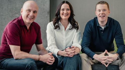 Martin Geissler, a bald man in a burgundy polo shirt and blue jeans, sits on a green sofa in a concrete-walled room. He is smiling. To his right Laura Maciver, a woman with long black hair, is wearing a white shirt and blue jeans and is also smiling. To her right sits Phil Goodlad. He is wearing a blue polo shirt and sand-coloured jeans and is also smiling. Copyright