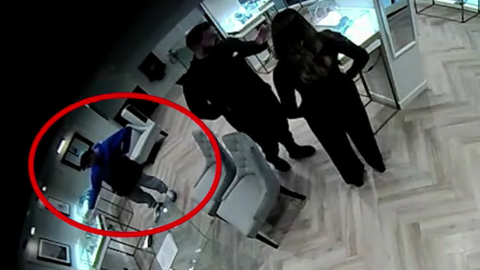 CCTV footage taken from a high angle inside a jewellery shop of a man talking to a female employee while another in the background puts his hand into a glass cabinet to steal jewellery