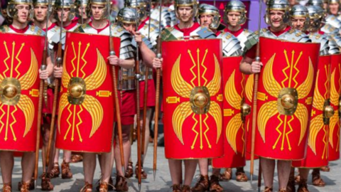 A large group of men dressed as ancient Roman legionaries. They are dressed in silver-coloured armour, including helmets, over red tunics. They are carrying spears and red shields with a yellow design on them. On their feet are leather sandals, tied up around their ankles. 