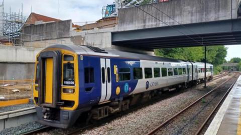 A blue, white and yellow Northern train passing through a station. It is made up of two carriages and there is a concrete bridge above it and building work behind it.