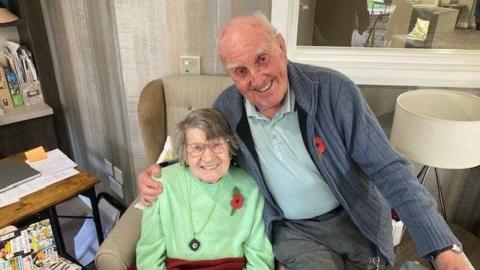 Michael Woods with his wife Mary. They are seated in an armchair with Michael perched on the arm. he gives his wife a hug. Both are smiling and wearing Remembrance poppies on their jumpers.