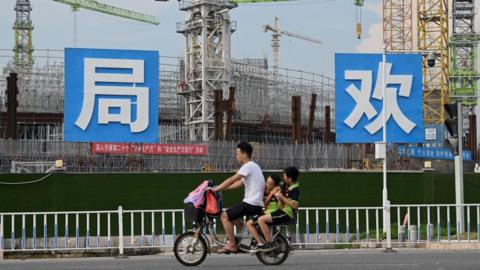 People commute in front of the under-construction Guangzhou Evergrande football stadium in Guangzhou, China's southern Guangdong province on September 17, 2021. The photo shows a male motorcyclist with two passengers, children, seated behind him, as they ride past a large construction site.