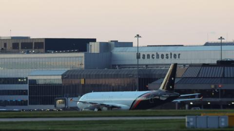 An airplane is on the runway, with grass in the foreground and an airport in the background with Manchester emblazoned on it