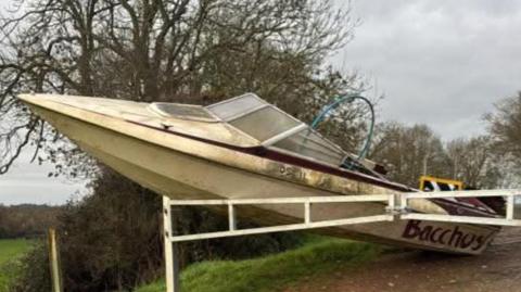 A cream coloured boat, shaped like a retro speedboat, leans against white barriers with its nose in the air, on a country lane leading into a field.