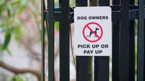 A white sign stating 'dog owners: pick up or pay up" can be seen close to black fence spikes. In the background, green leaves can be made out. 