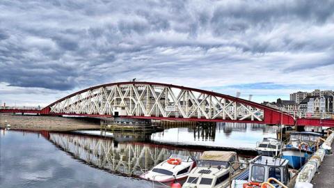 The Ramsey Swing bridge, viewed from across the harbour, is a red arched structure with white metal columns and diagonal metal support beams. Several boats moored near the bridge in Ramsey harbour.