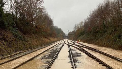 Murky flood water on railway tracks. There are trees and bushes on either side of the lines.