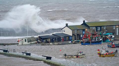 Elevated view of buildings on the Cobb harbour wall in Lyme Regis, now protected by a second breakwater in the background. The sea is grey and waves are crashing against the newer outer wall. Four small fishing boats are anchored inside the harbour in the foreground where the water is choppy but comparatively calm.