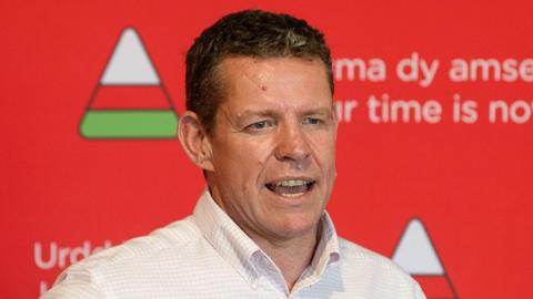 Rhun ap Iorwerth is wearing a white shirt and a rosette, in front of the red Urdd Eisteddfod branding.