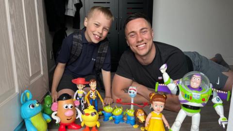 A child and his father lying at a doorstep with an array of toys from the Toy Story film franchise