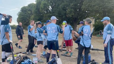 London Softball League team MI6 are gathered around amongst bats and other softball equipment on the grassy floor. Players, both male and female, are wearing light-blue softball t-shirts. Some are wearing dark-blue shorts, others are wearing jogging bottoms. One player, bearing the number 25 on the back of her shirt, is balancing a green softball on the back of her hand. 