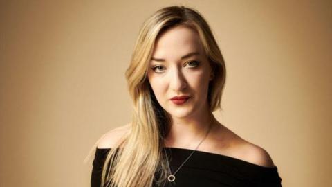 Woman with long blonde hair and dark top, wearing a necklace, looking at camera.