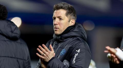 QPR boss Julien Stephan, wearing a navy blue coat with the QPR badge on it, is clapping.