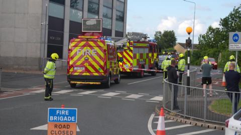 Three red and yellow fire service vehicles sits in a line outside a grey bricked building. They have cordoned off the area using red and white tape. Four fire men in illuminous yellow jackets can be seen beyond the cordon with one in front of it. A gathering of three men watch on from in-front of the cordon. A blue sign in white font reads 'Fire Brigade'. An orange sign in black font reads 'Road Closed.'