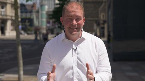 Wales Business Correspondent, Huw Thomas, is seen looking into the camera, wearing a white shirt. The background is a blurry high street.