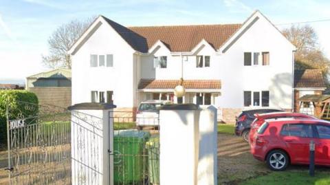 A whitewashed building with metal gates in front of it. There a four cars parked in the driveway