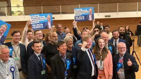 Paul Bristow celebrates with his supporters in the counting centre hall. Some of the group are holding blue Conservative Party placards and others are wearing blue Conservative Party rosettes. 