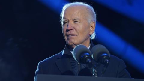 Joe Biden stands at a lecturn wearing an overcoat, he is looking off to his right and looks to be slightly squinting 