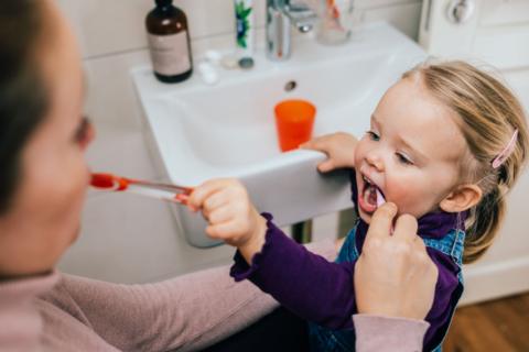 A little girl is standing by a sink with a woman standing next to her. They are brushing each other's teeth. 