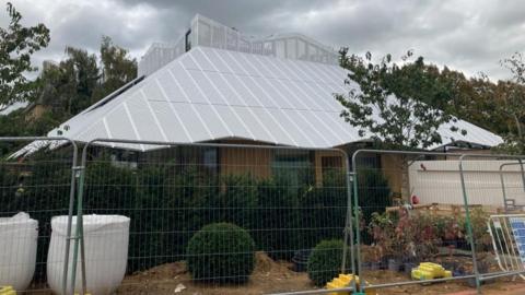 A building behind fencing. The roof is grey and shaped like a hut. There are trees in front.