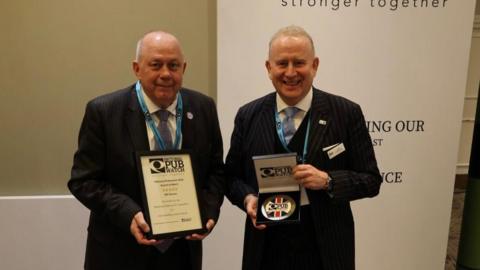 Two grey-haired men wearing suits and smiling at the camera. The one on the left is holding up a certificate and the one on the right is holding a plaque. Both items have the National Pubwatch logo on them