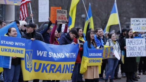 Protesters holding Ukraine flags and pro-Ukraine signs with the words "We Stand With Ukraine", "Ukraine Strong" and "America Stands With Ukraine"