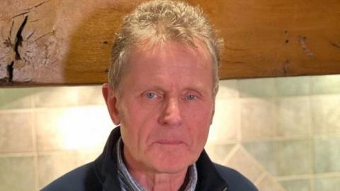 Warwick Jackson has short grey hair. He is wearing a navy blue zip-up jumper with a blue striped shirt underneath. He is standing in front of a blue tiled wall and a wooden shelf.