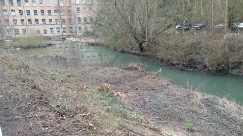 Area of scruffy, cleared scrub land in a dip, with a strip of water at the bottom, leading up to a historic mill building